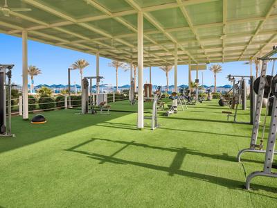 Espace de gym extérieur couvert avec divers appareils d'exercice et vue sur des palmiers.