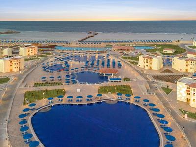 Groot hotelzwembad met veel blauwe parasols en uitzicht op de zee en haven.