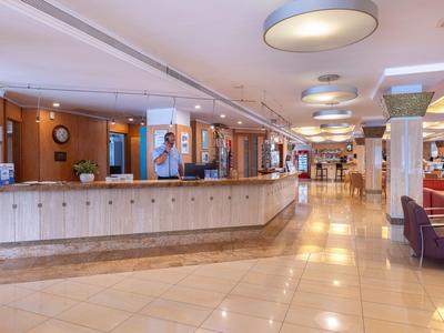 Helle Hotellobby mit langer Rezeption, runden Deckenleuchten und Sitzbereich mit roten und lila Stühlen.