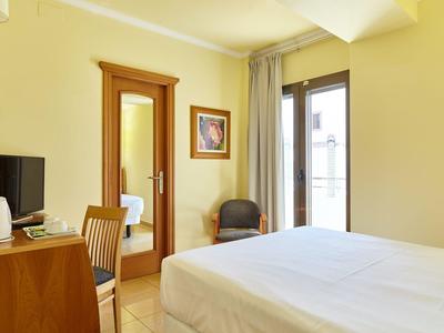 Helles Hotelzimmer mit Doppelbett, Schreibtisch, Fernseher und Balkonfenster.