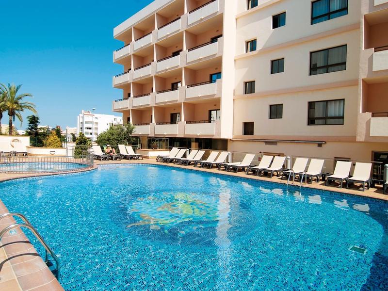 Outdoor swimming pool surrounded by lounge chairs and a multi-story building under clear blue sky.