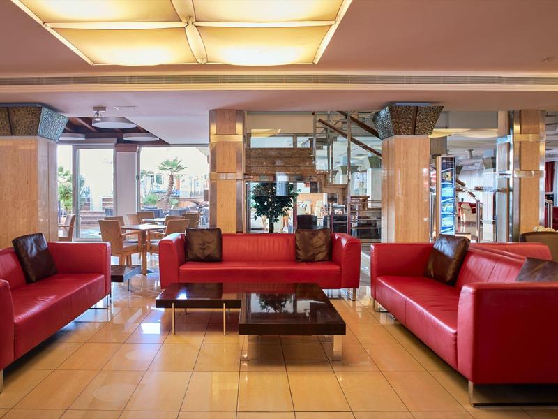 Hotel lobby with red leather sofas, large windows, and a glass coffee table on tiled floor.