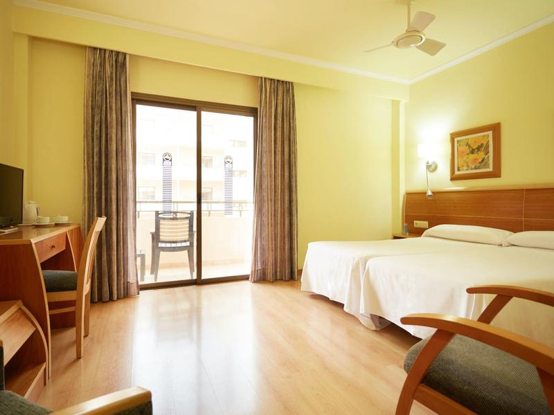 Bright hotel room with twin beds, wooden floor, desk, TV, and balcony with chair.