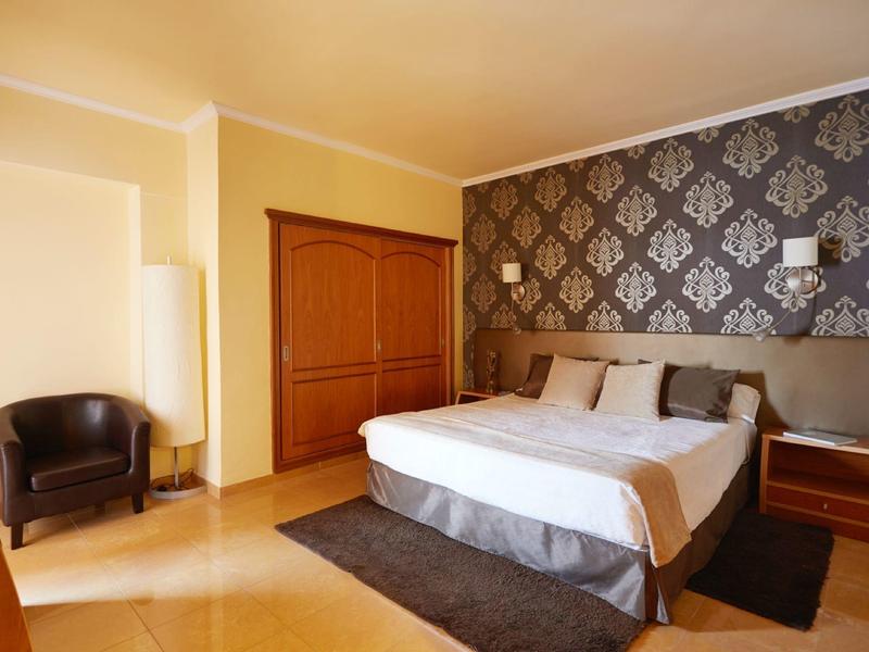Hotel room with double bed, patterned wallpaper, armchair, and wooden closet.