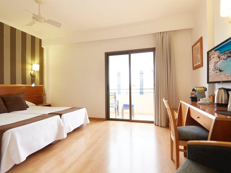 Bright hotel room with wooden floor, two beds, desk, chair, and balcony door with curtains.
