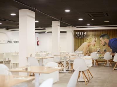 Moderní, prostorná kavárna s dřevěnými stoly, bílými židlemi a žlutou pruhovanou zdí s nástěnnou malbou.