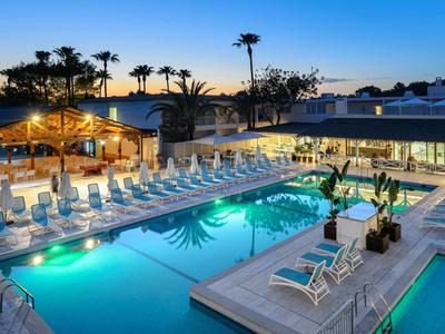 Moderner Außenpool mit Liegestühlen, beleuchtet bei Sonnenuntergang, umgeben von Palmen und Gebäuden.