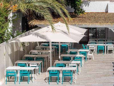 Venkovní jídelní prostor s bílými stoly, tyrkysovými židlemi, zastíněný velkým bílým slunečníkem u palem.