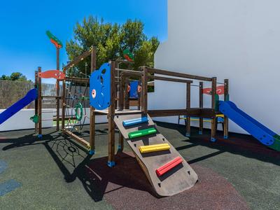 Kinder-Spielplatz mit Klettergerüst, Rutschen und hölzernen Strukturen neben einem weißen Gebäude.