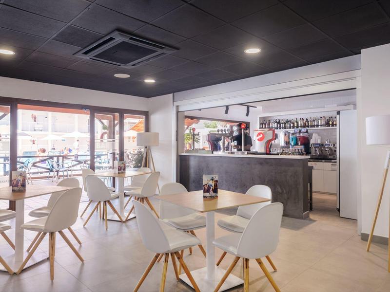 Moderní hotelový bar s bílými židlemi, dřevěnými stoly a lesklým černým pultem pod tmavým stropem.