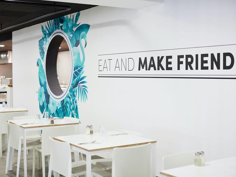 Bílá jídelna s stoly, židlemi a nápisem na zdi 'JEDZTE A NAHRAJTE PŘÁTELE'