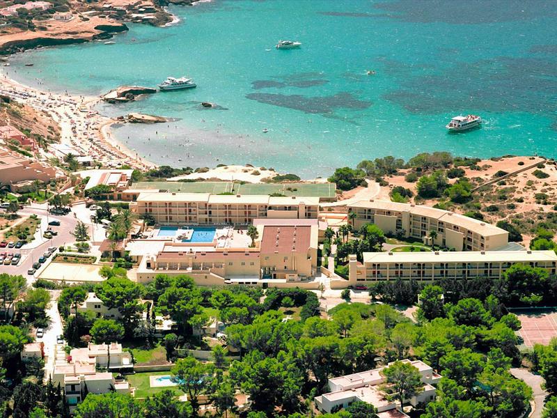 Pobřežní hotelový resort s bazénem, obklopený zelení a poblíž tyrkysového moře s loděmi.