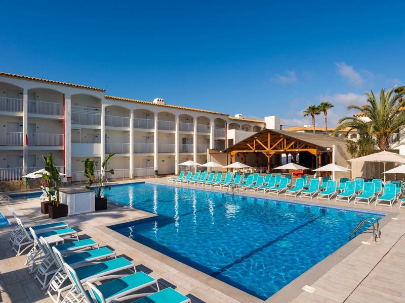 Venkovní hotelový bazén s modrými lehátky a jasnou oblohou.