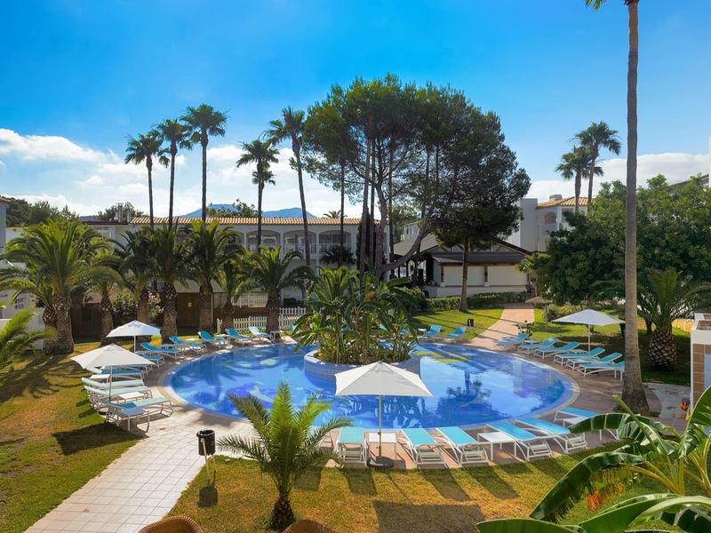 Venkovní hotelový bazén obklopený lehátky, slunečníky, palmami a bujnou zelení pod modrou oblohou.