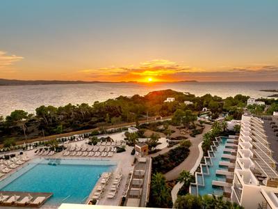 Hotel mit Pool und Meerblick bei Sonnenuntergang, mit Liegestühlen und grünen Bäumen.