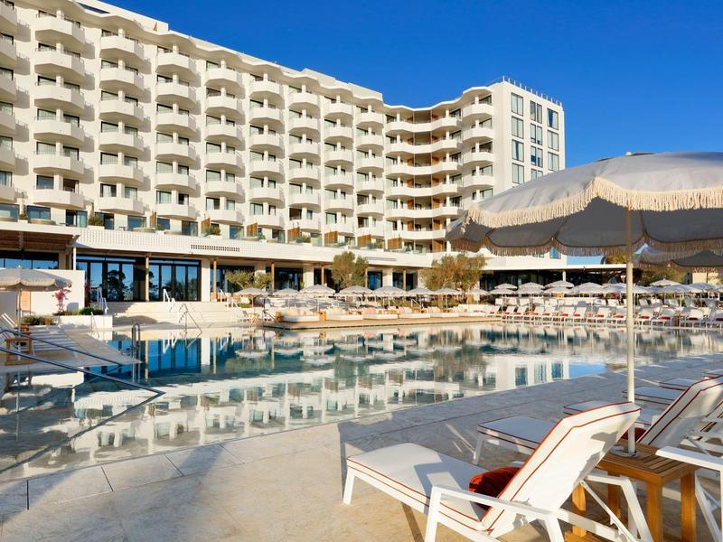 Hotel s venkovním bazénem, lehátky a béžovo-bílými slunečníky pod modrou oblohou.