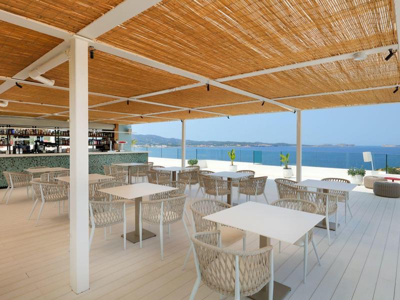 Otevřená terasa s bílými stoly a židlemi, v pozadí výhled na moře.