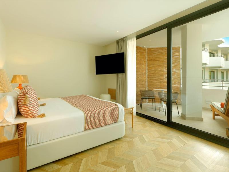 Světle hotelový pokoj s manželskou postelí, TV a balkonem se židlemi a výhledem.