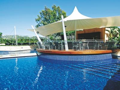 Espace piscine moderne avec eau bleue, voiles d'ombrage et terrasse en bois dans un cadre ensoleillé.