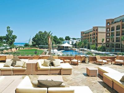 Cour avec canapés lounge, vue sur jardin, piscine et bâtiment hôtelier à plusieurs étages sous ciel bleu.