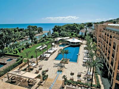 Complexe hôtelier avec piscine, chaises longues, palmiers et vue sur la mer sous un ciel dégagé.