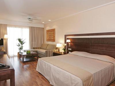 Chambre d'hôtel lumineuse avec grand lit double, canapé, bureau et sol en bois.