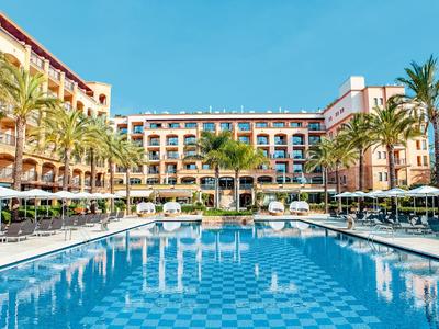 Großes Hotel mit Außenpool, Sonnenliegen und Palmen unter klarem Himmel.
