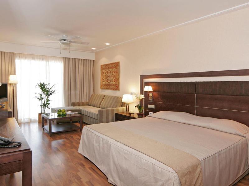 Chambre d'hôtel lumineuse avec grand lit double, canapé, bureau et sol en bois.