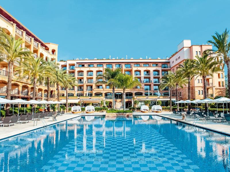 Hôtel de luxe avec grande piscine, palmiers et parasols sous un ciel dégagé.