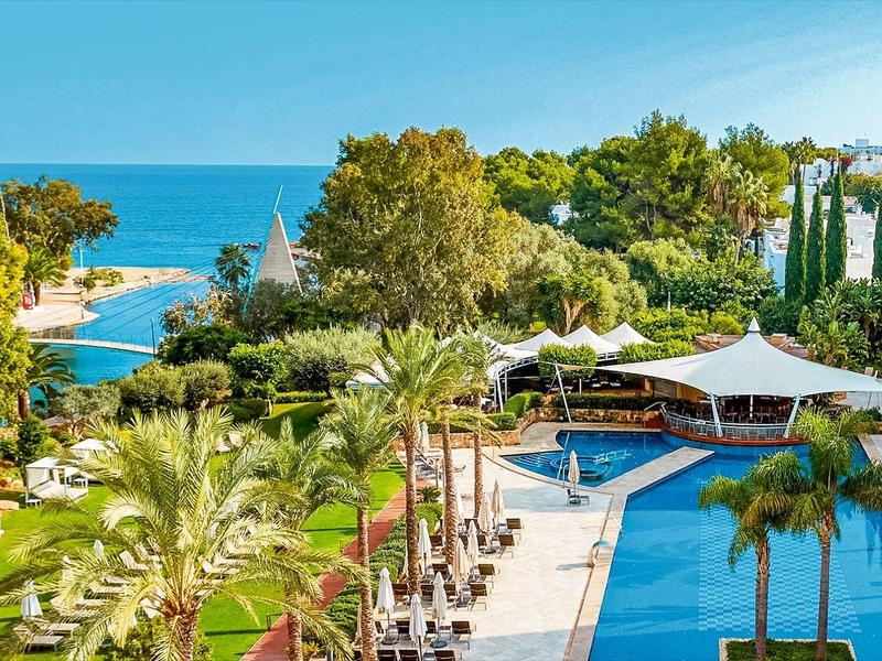 Complexe hôtelier avec piscine, palmiers et vue directe sur la mer sous un ciel dégagé.