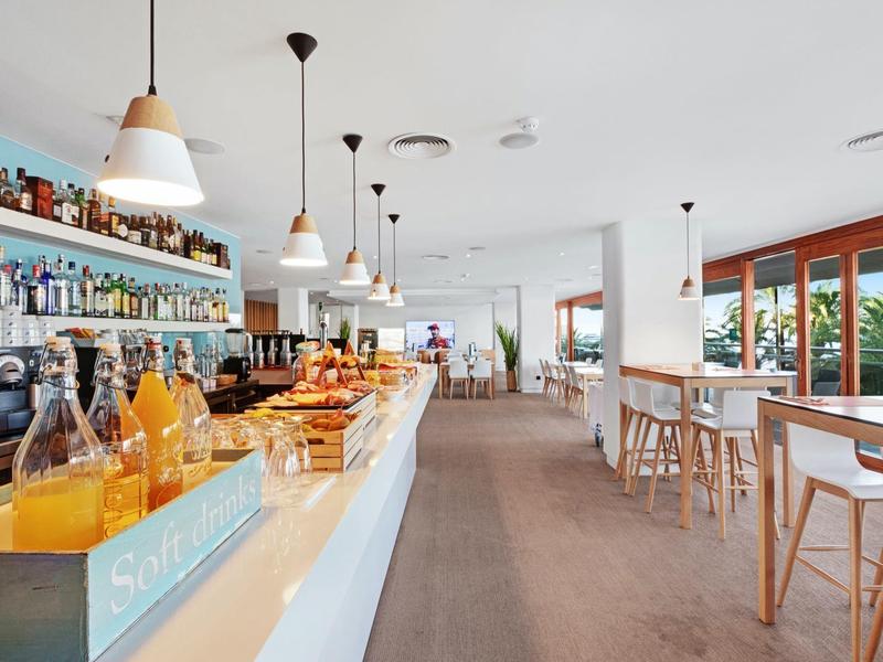 Moderní světlý bar s nápoji, pečivem a vysokými židlemi u velkých oken.