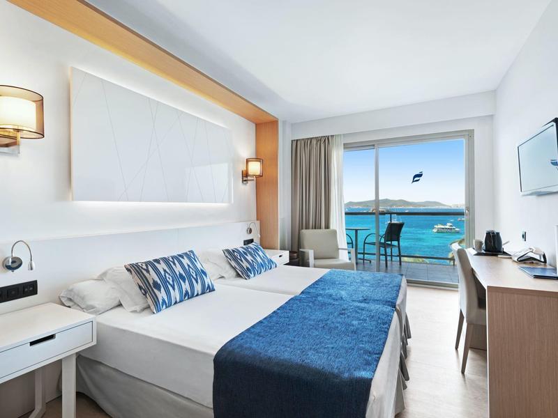 Moderní hotelový pokoj s výhledem na moře, manželskou postelí, balkonem a modrobílým zařízením.