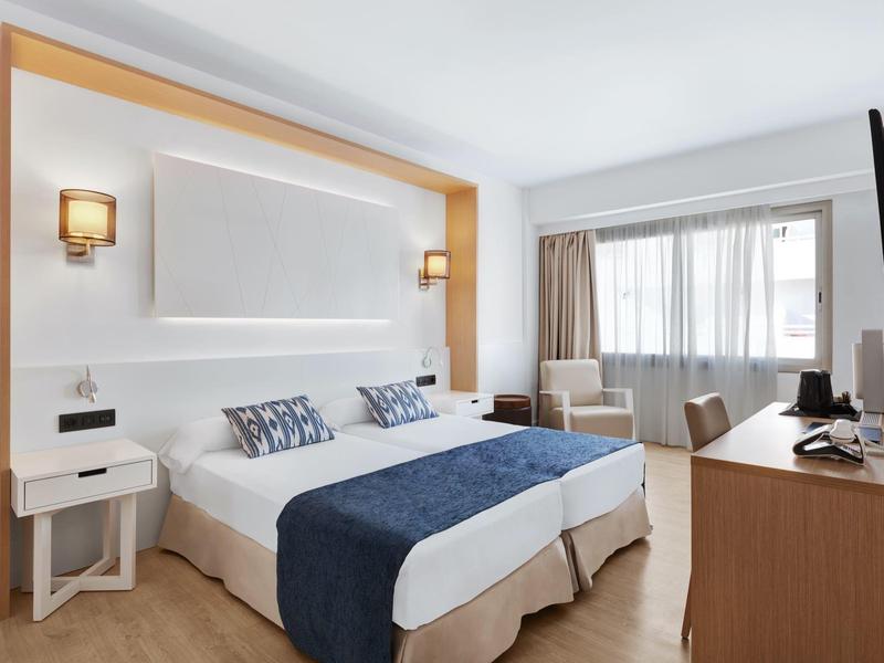 Moderní hotelový pokoj s manželskou postelí, televizí na zdi a velkými světlými okny.