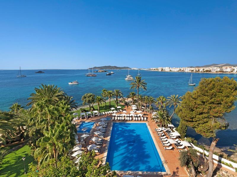 Modrý bazén obklopený palmami, výhled na moře a jasnou oblohu.