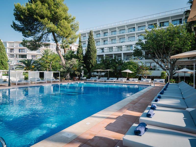 Venkovní bazén s lehátky vedle moderního hotelu obklopeného stromy.