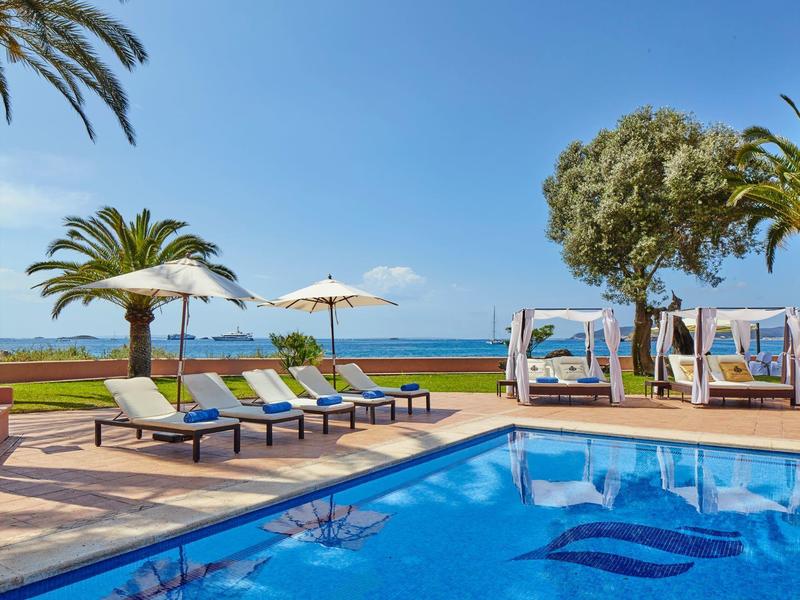 Světle modrý bazén s lehátky a slunečníky s výhledem na moře za jasné oblohy.