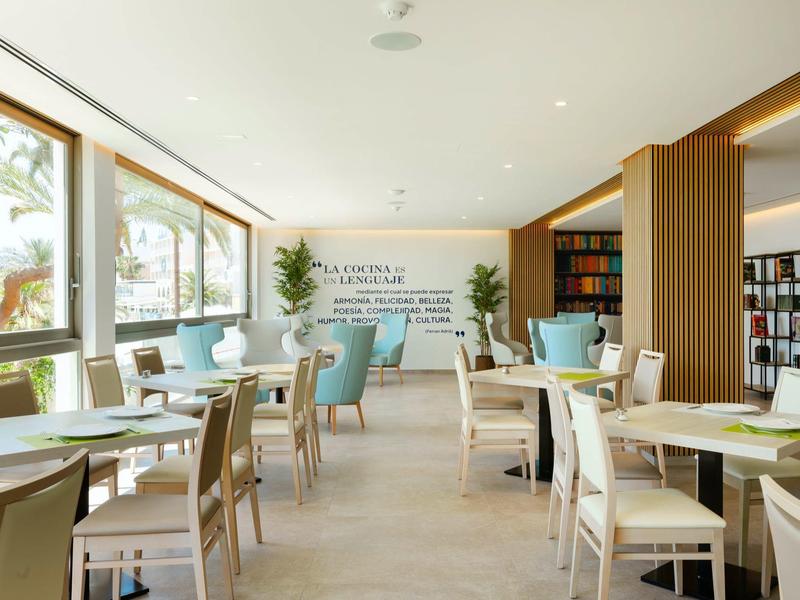 Jasná jídelna s velkými okny, dřevěnými stoly a barevnými židlemi.