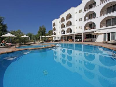 Large blue swimming pool in front of a white hotel building with balconies and umbrellas.