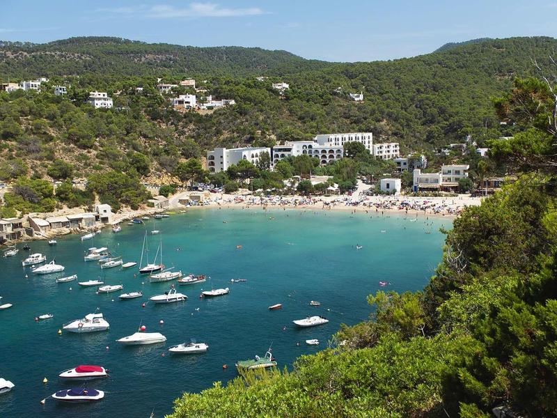 Coastal bay with clear water, boats, and hotels surrounded by green hills.