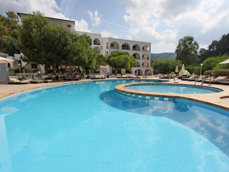 Large outdoor pool with sun umbrellas and hotel building in the background under cloudy sky.
