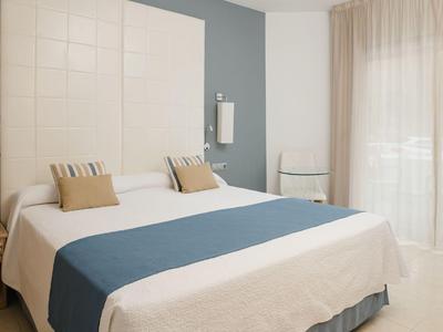 Hell eingerichtetes Hotelzimmer mit großem Bett, blauer Tagesdecke und großem Fenster mit Vorhängen.