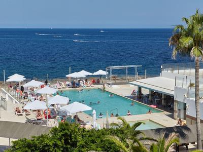 Area piscina dell'hotel con ombrelloni bianchi e vista sul mare blu.