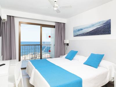 Nowoczesny pokój hotelowy z łóżkiem podwójnym, niebieskimi akcentami i widokiem na morze przez szklaną ścianę.