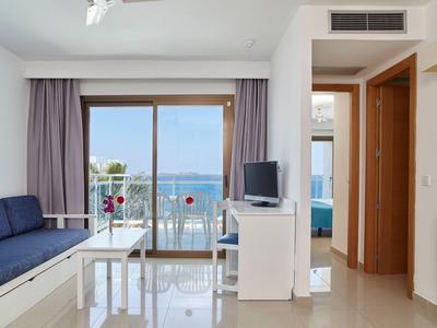 Jasny salon z ciemnoniebieską kanapą, białym stołem, telewizorem i widokiem na morze przez duże okno.