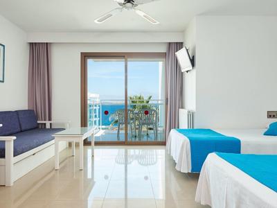 Pokój hotelowy z dwoma łóżkami, sofą i widokiem na morze przez duże szklane drzwi przesuwne.