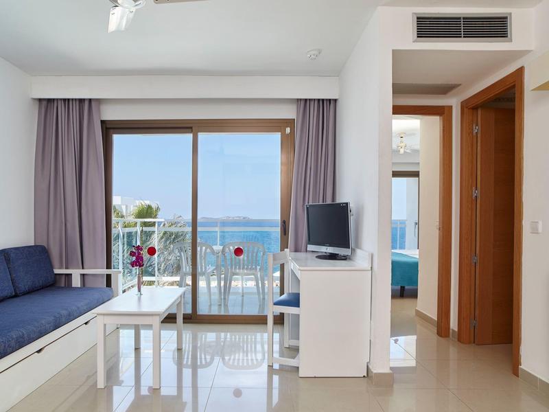 Jasny salon z ciemnoniebieską kanapą, białym stołem, telewizorem i widokiem na morze przez duże okno.