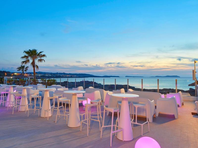 Taras z białymi krzesłami i stołami o zachodzie słońca z widokiem na morze i palmy.