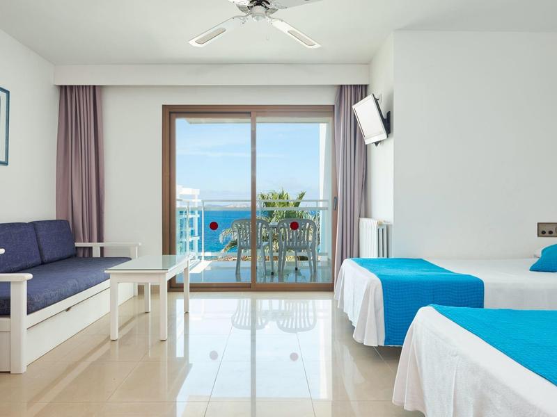 Pokój hotelowy z dwoma łóżkami, sofą i widokiem na morze przez duże szklane drzwi przesuwne.