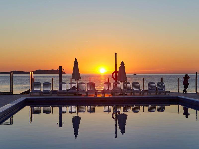 Zachód słońca nad basenem z leżakami i parasolami przy wodzie.