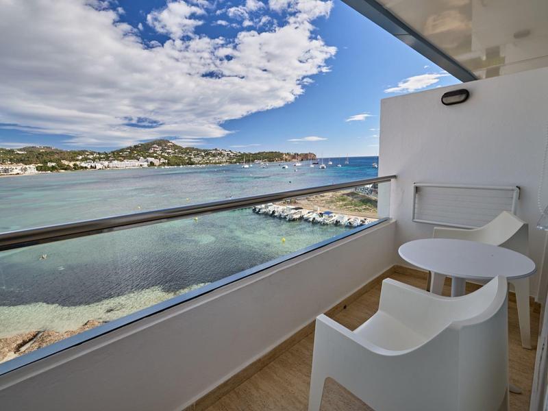 Balcony with white chairs and table overlooking the sea and beach under a blue sky.